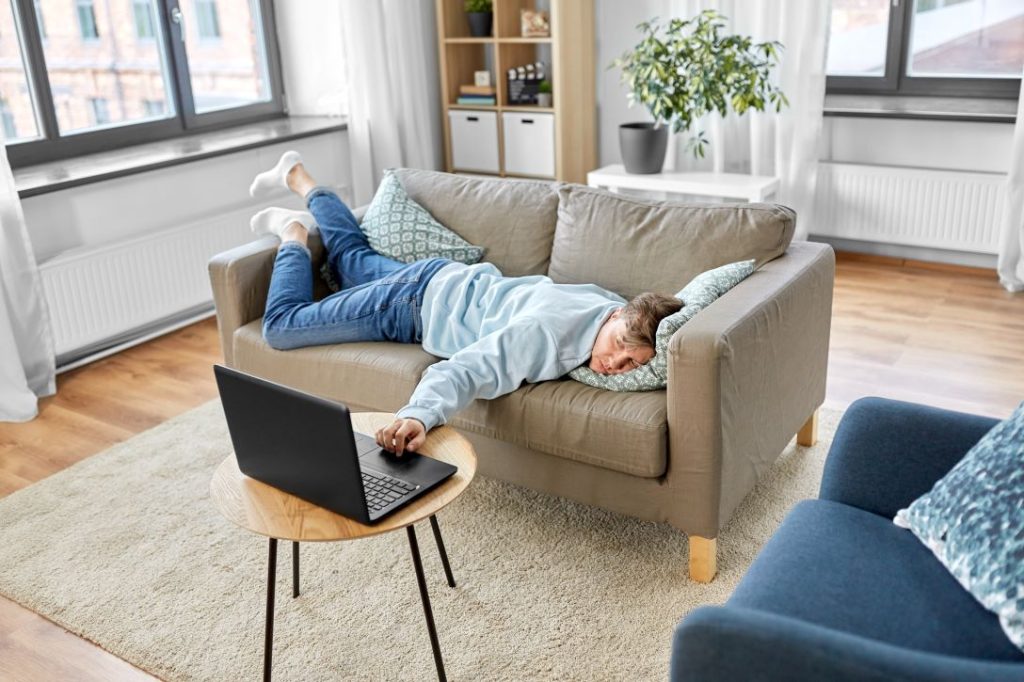 Man laying facedown on couch with arm stretched out to a laptop