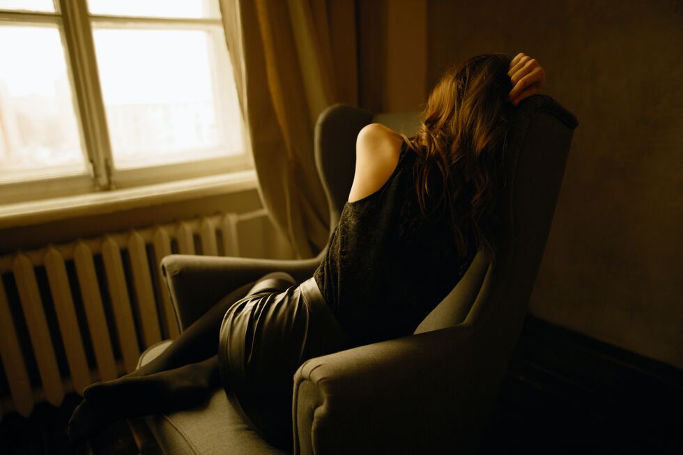 woman in dark clothes on dark couch, window looking outside, beige curtains